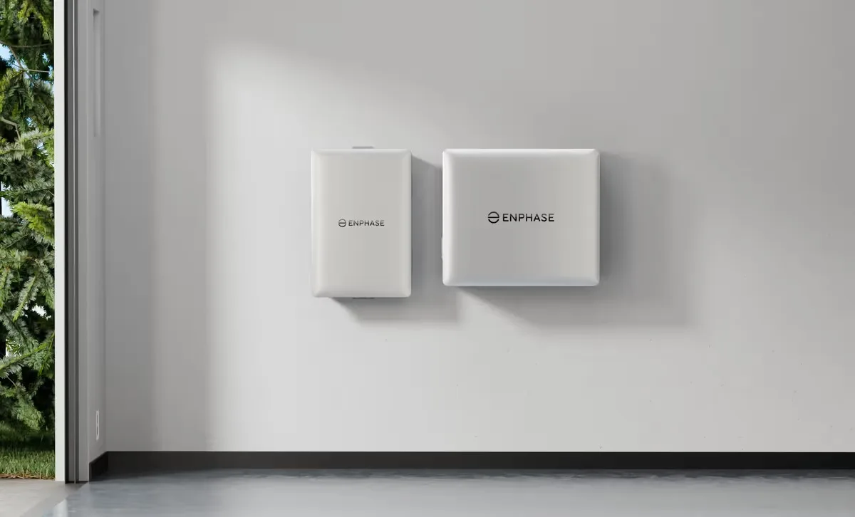 Two white Enphase home batteries mounted on a garage wall next to an open garage door showing trees.