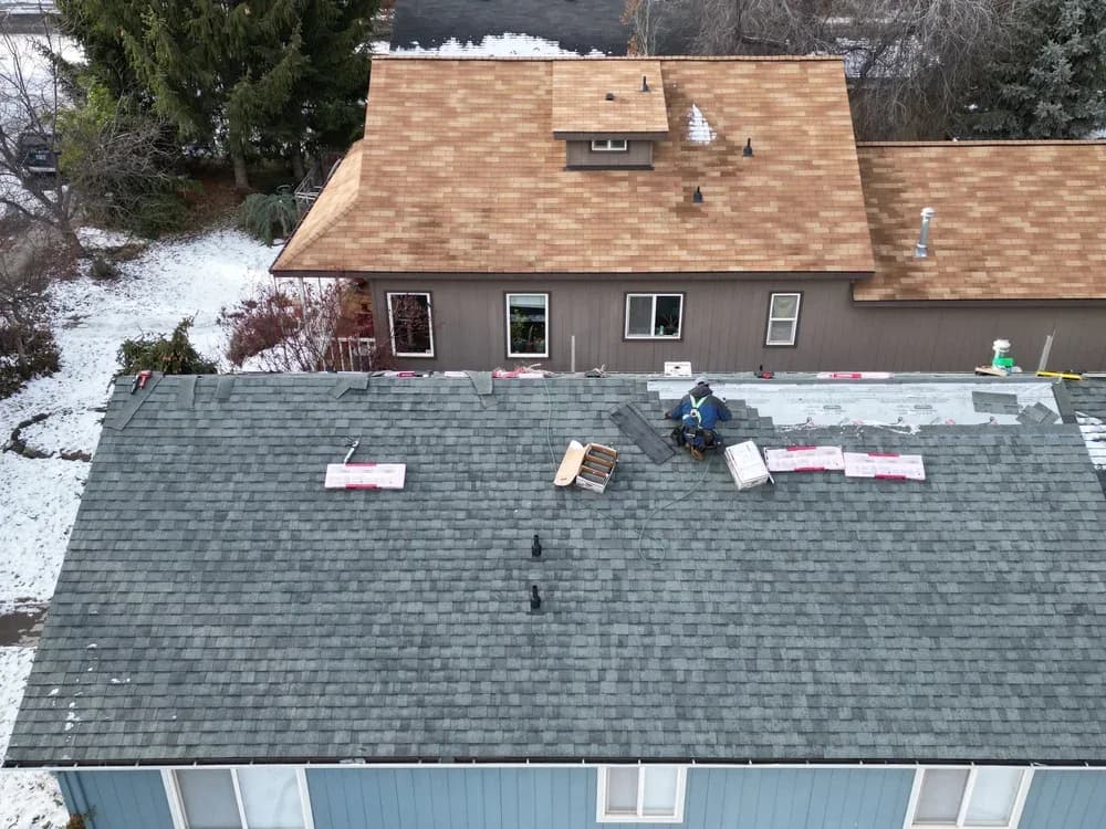 Roofer installing asphalt shingles on a snow-dusted residential roof with stacked materials and adjacent house