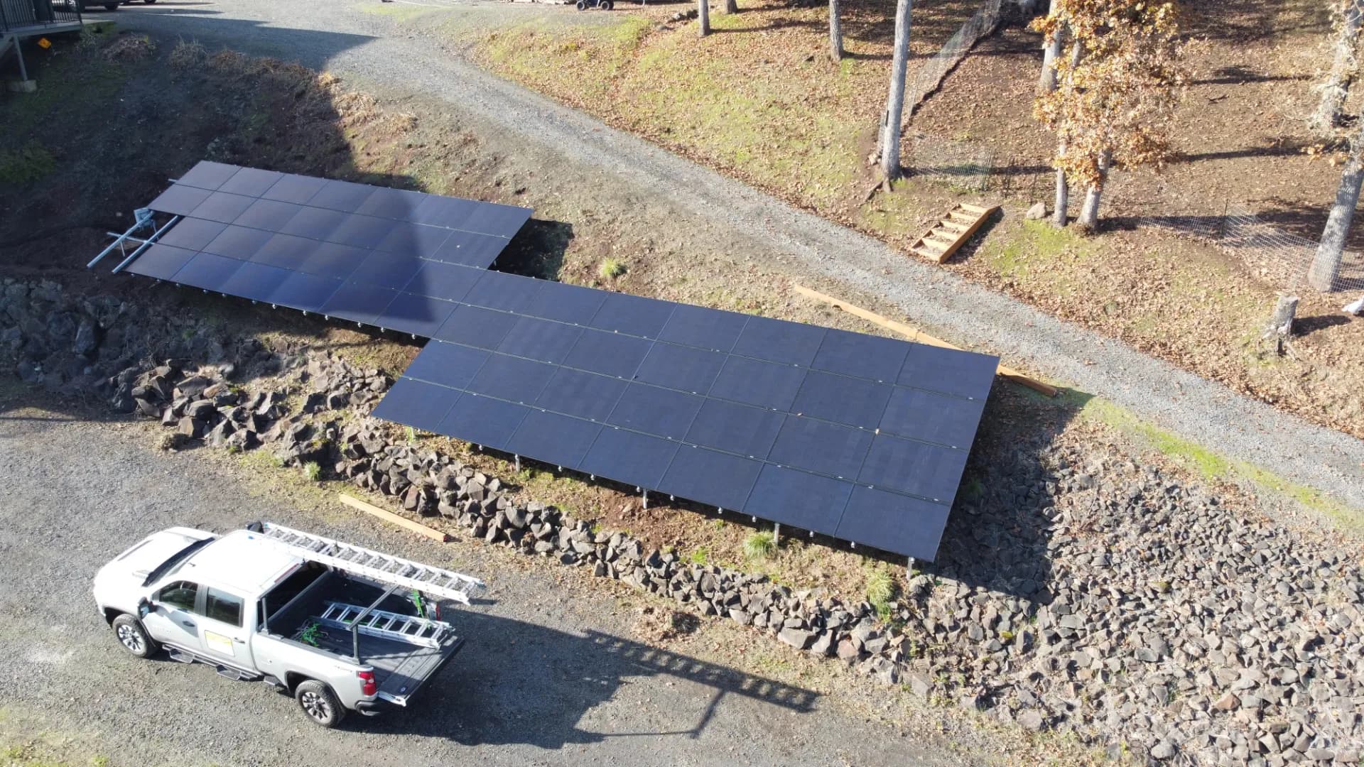 Ground-mounted solar array on a rocky slope beside a gravel drive, service pickup with ladder parked in foreground.