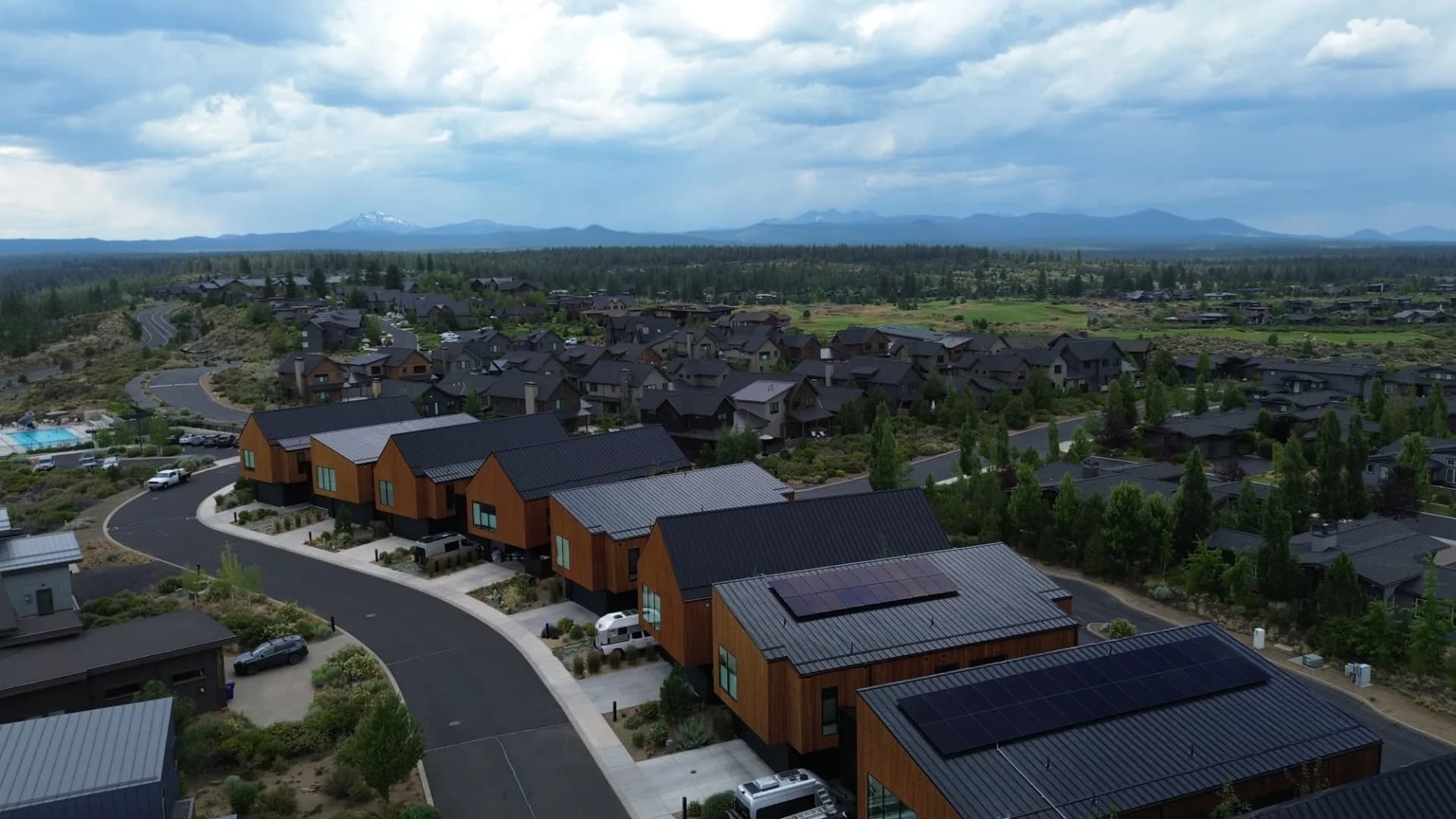 Aerial view of Oregon neighborhood with homes, metal roofs and rooftop solar panels, mountains and cloudy sky.