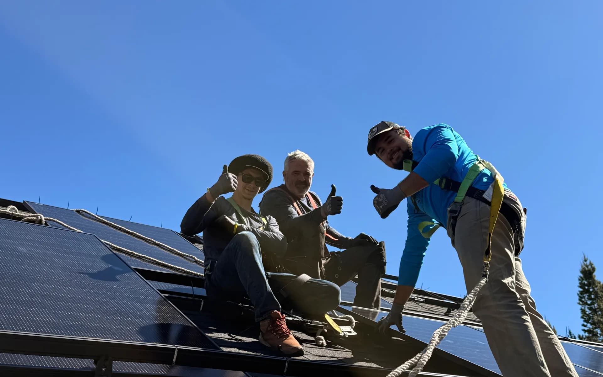 Three rooftop solar installers wearing harnesses give thumbs-up while sitting on solar panels under a clear blue sky