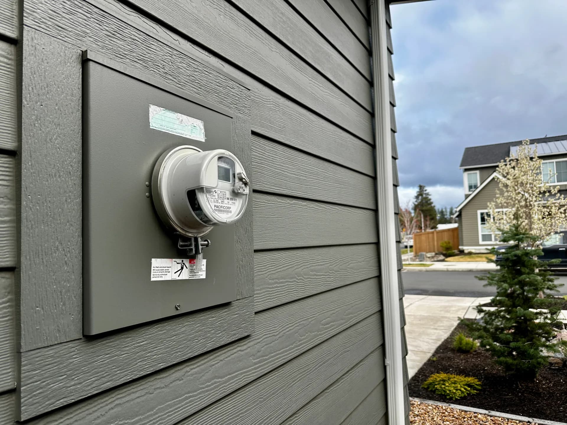 Electric utility meter mounted on gray house siding, neighboring homes and cloudy sky in the background