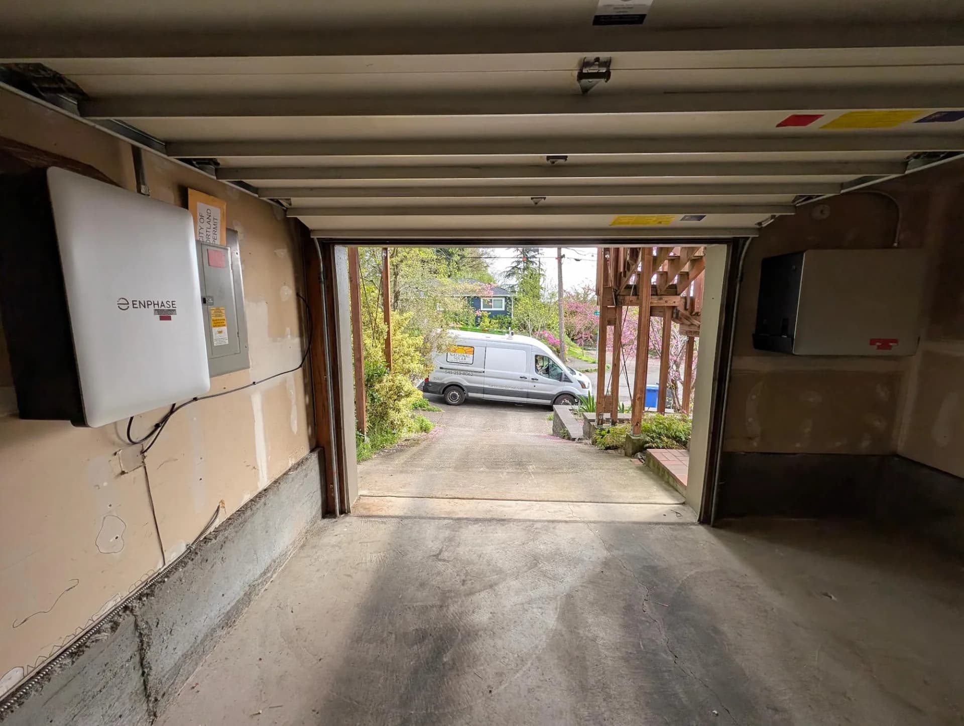 Open garage facing driveway, Enphase solar inverter and battery enclosures mounted on walls, service van outside.