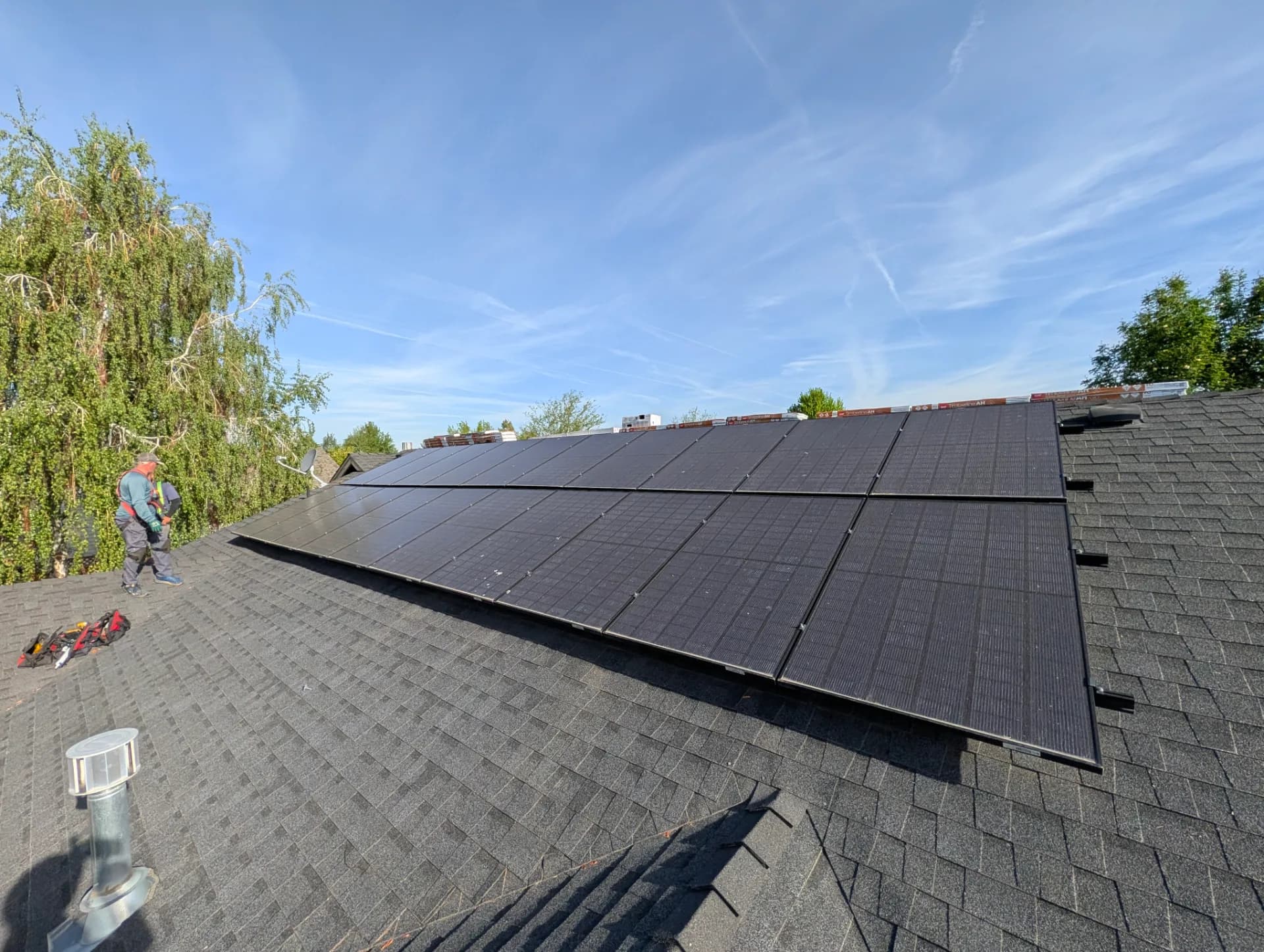 Rooftop solar array on asphalt-shingle roof with installers and tools beside trees under a clear blue sky.