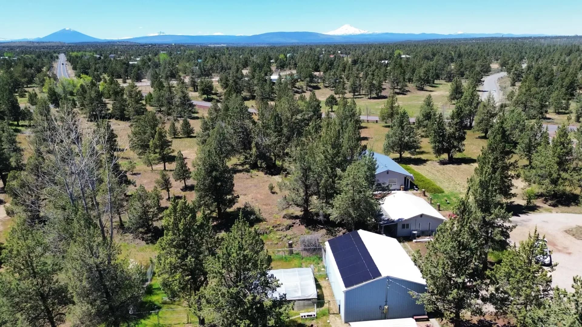 Aerial view of rural Bend property with rooftop solar panels, pine trees, winding roads and snow-capped Cascade peaks.